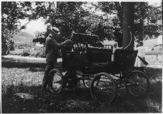 Man behind early automobile, 1890s?, women in background, umbrella, trees, field   Prints